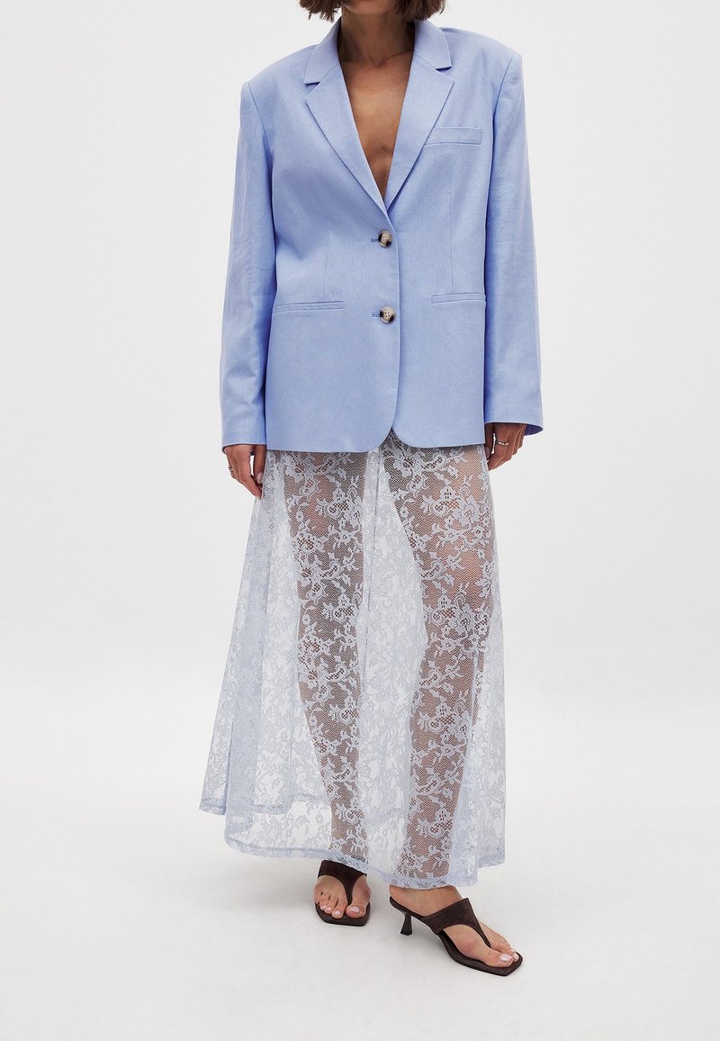 Svetlo modra lanena blazerica z dvema gumboma, kombinirana z prosojno belo čipkastno krilo in črnimi čevlji s peto; teksturirana tkanina in eleganten vzorec.