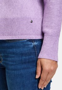 Hellblau gerippter Pullover mit strukturiertem Oberteil und kontrastierenden gerippten Manschetten, mit einem kleinen metallischen Akzent nahe dem Saum. Kombiniert mit blauen Jeans.