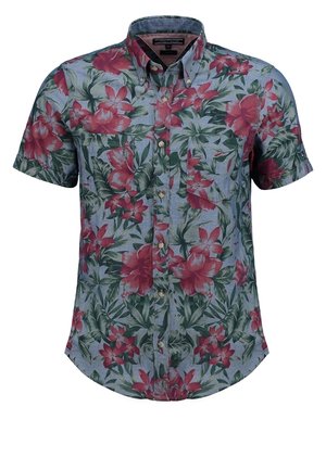 Mężczyzna w krótkim, kwiecistym koszulowym t-shircie, ciemnych, podwiniętych dżinsach i brązowych skórzanych butach stoi na jednolitym tle.