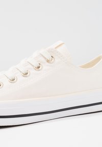 Baskets en toile couleur blanc cassé avec bout rond, six œillets métalliques et lacets blancs. Présente une bande noire traversant la semelle en caoutchouc blanche.