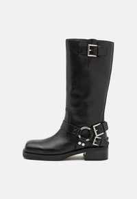 Botte noire en cuir montant jusqu'au genou avec des boucles en argent et un accent circulaire, dotée d'un bout arrondi et d'un petit talon carré. Texture lisse.