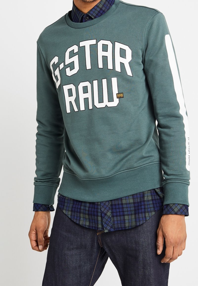 Grön sweatshirt med stora vita "G-STAR RAW" bokstäver, ribbade ärmslut och nederkant, med ett kontrasterande vitt rand på ärmen och en lager-på-lager rutiga skjorta under.