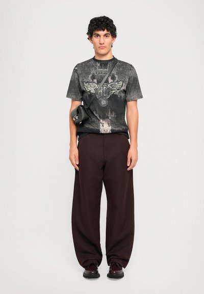 Man wearing black and gray graphic t-shirt, dark brown wide-leg pants, black crossbody bag, and dark shoes standing against white background.