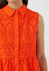 Oranssi ilman hihoja oleva mekko napituksella edessä, jossa on monimutkaisia kirjontakuvioita ja rei'itettyjä yksityiskohtia sekä kerrottu hame.