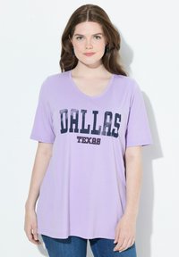 Mujer con cabello castaño lleva una camiseta V de color lavanda con el texto "DALLAS TEXAS" y jeans azules, de pie contra un fondo blanco.