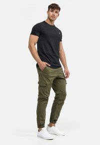 Tricou negru cu mâneci scurte, pantaloni cargo verde măsliniu cu manșete elastice și teniși albi; are buzunare laterale și un croi strâns.
