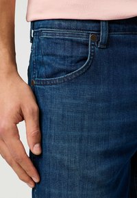 Jeans de mezclilla azul oscuro con una superficie suave, un ligero desgaste, detalles en los bolsillos delanteros y acentos de metal en bronce.