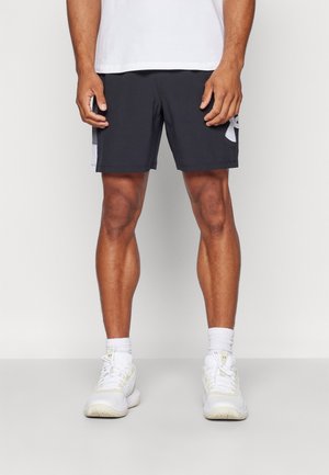 Homme portant un short de sport noir avec un logo blanc sur la cuisse droite, des chaussettes blanches montantes, des baskets blanches, debout devant un fond uni.