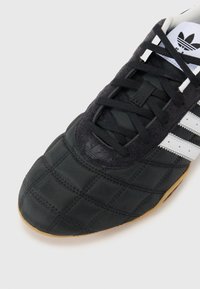 Black athletic shoe featuring a quilted design, suede accents, white stripes, and a rubber sole. Laces are black and securely fastened.