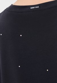Zwart T-shirt met een ronde hals, voorzien van kleine, glanzende versieringen verspreid over de rug. Label zichtbaar bij de hals.