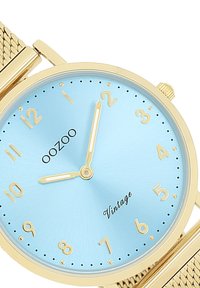 Montre-bracelet dorée avec un cadran bleu clair, des index et des aiguilles dorés. Dispose d'un bracelet en maille texturée. Nom de la marque 'OOZOO' affiché.