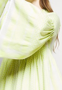 Světlé zelené šaty s nařasenými rukávy a texturovanou látkou s vzory teček a čar. Měkký, splývavý design dodává objem.