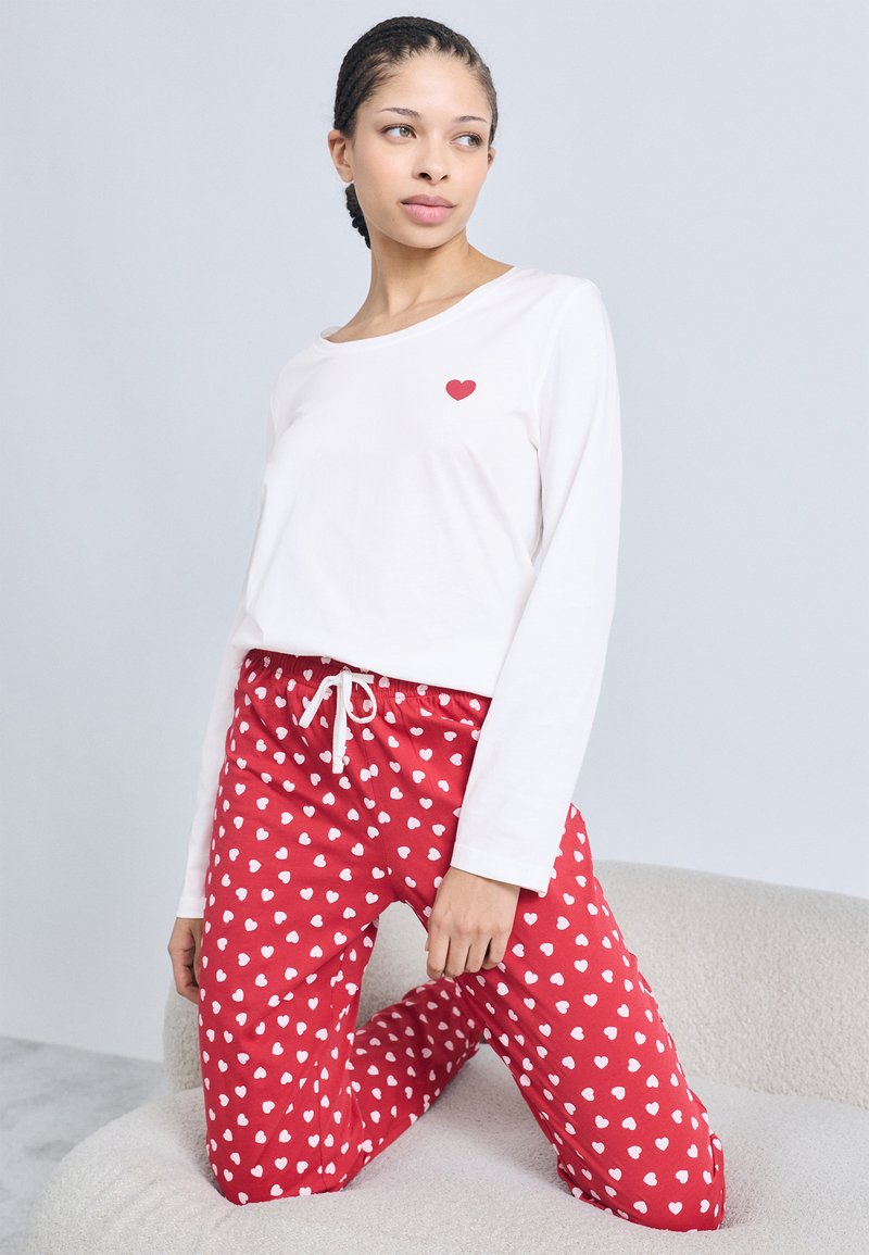 Άσπρο μακρυμάνικο τοπ με κόκκινη καρδιά, σε συνδυασμό με κόκκινη φόρμα με μικρές άσπρες καρδιές και ρυθμιζόμενο κορδόνι στη μέση.