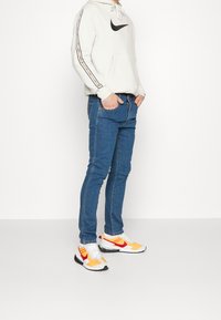 Man som bär en vit Nike-hoodie, blå slim jeans och vit-orange Nike-sneakers, står med händerna i fickorna mot en helt vit bakgrund.