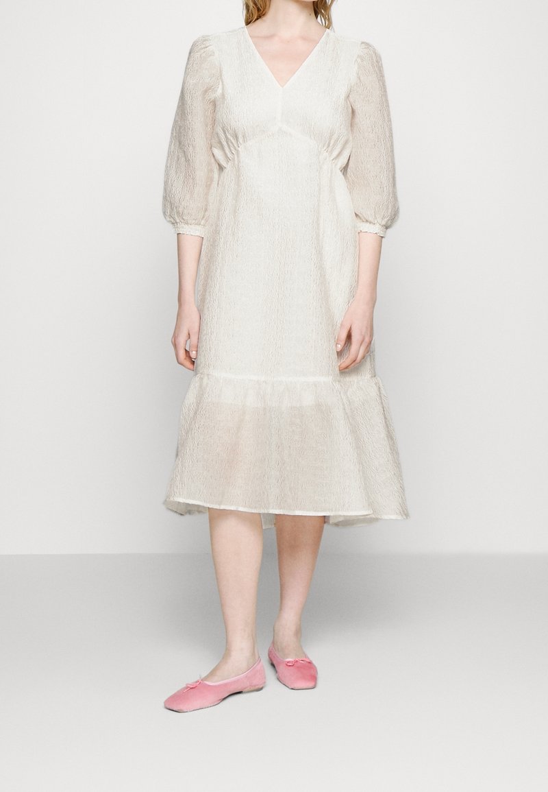 Robe blanche texturée avec un col en V, des manches bouffantes et un ourlet à volants, associée à des chaussures plates rose pâle.