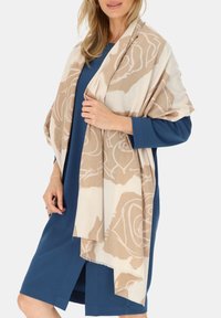 Beżowy szalik z kwiatowym wzorem, wykonany z miękkiego materiału. Przerzucony na niebieską sukienkę, z frędzlami na brzegach i luźnym, swobodnym krojem.