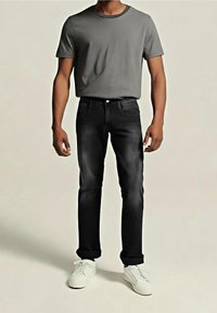 Homme portant un t-shirt gris à manches courtes, un jean noir avec des revers et des baskets blanches, debout devant un fond clair uni.