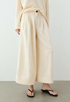 Personne portant un pantalon ample couleur crème à jambes larges avec un bouton noir et des sandales à talons noires, debout sur un sol gris devant un mur uni.