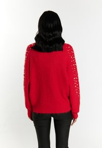 Maglione rosso lavorato a maglia con una vestibilità ampia, superficie testurizzata e decorazioni di piccole perle sulle spalle. Indossato con pantaloni neri aderenti.
