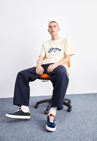 Camiseta crema con logo azul, pantalones oscuros de corte holgado y zapatillas multicollor; sentado en una silla de oficina naranja frente a un fondo claro.