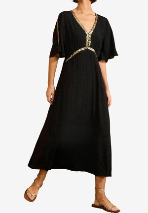 Vrouw met een zwarte midi-jurk met gouden paillettenafwerking op de V-hals, taille en mouwen, gecombineerd met gouden sandalen met fijne bandjes en een platte zool.