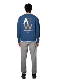 Niebieska bluza z dużym białym i złotym napisem z tyłu, zestawiona z szarymi dresami, noszona z czarnymi butami. Swobodny strój sportowy.