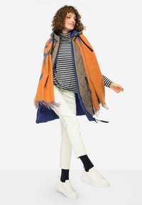 Gilet matelassé gris avec capuche, enveloppé dans une couverture orange avec des franges, haut rayé noir et blanc, pantalons blancs et baskets blanches.