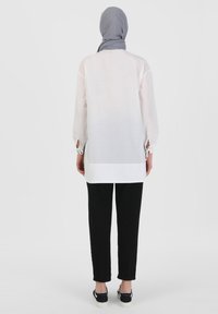 Lange witte blouse met ballonmouwen, open zijslitsen en strikaccenten. Gepaard met slanke zwarte broek en instap schoenen. Grijze hoofddoek.