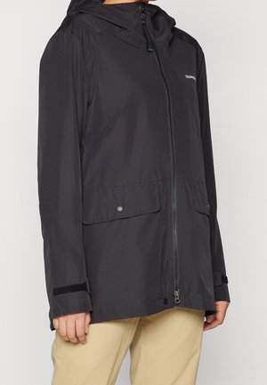 Schwarze Kapuzen-Regenjacke mit Frontreißverschluss, Klappentaschen und verstellbaren Handgelenkriemen, getragen mit beigen Hosen.