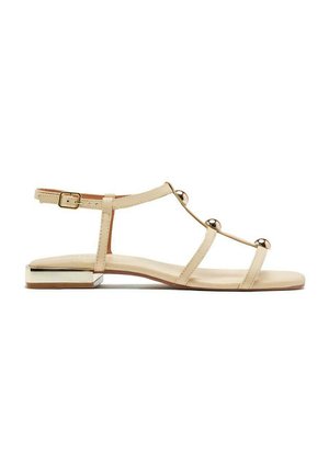 Sandalias planas beige con tres cintas finas y acentos metálicos redondos. Cuentan con una correa en el tobillo con hebilla y un tacón bajo cuadrado con un borde metálico.