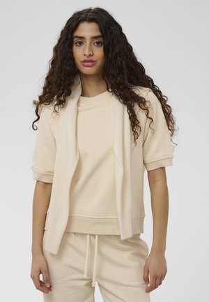 Femme aux longs cheveux bouclés portant un pull beige à manches courtes et un pantalon assorti, avec un pull posé sur ses épaules.