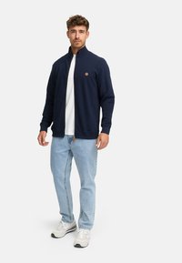 Hombre con chaqueta azul marino con cremallera, camiseta blanca, jeans claros y zapatillas beiges, de pie contra un fondo claro y simple.