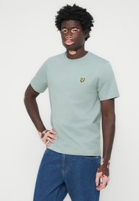 T-shirt en coton vert clair avec un col rond, des manches courtes et un petit logo jaune sur la poitrine. Porté avec un jean en denim bleu.