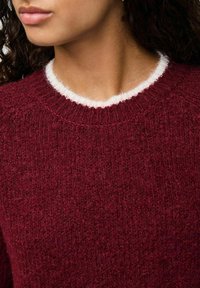 Femme portant un pull en maille rouge foncé texturé avec une bordure blanche duveteuse autour du col rond, vu de la mâchoire à la poitrine.