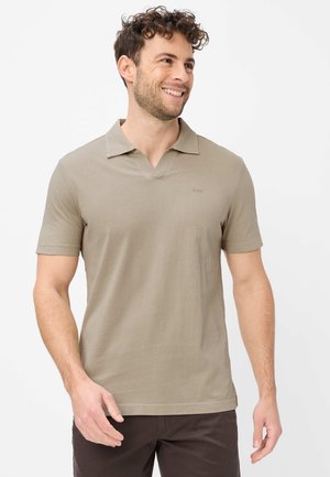 Smilende mand med krøllet hår iført en beige kortærmet poloshirt og mørkebrune bukser, stående mod en lys baggrund.