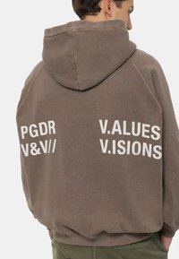 Felpa cappuccio marrone con testo stampato bianco "PGDR V&V// V.ALUES V.ISIONS" sulla schiena. Presenta una vestibilità rilassata e una texture morbida.