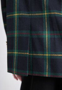 Blazer en mélange de laine avec un fond bleu marine foncé orné d'un motif à carreaux verts, jaunes et bordeaux, et doté d'une fente latérale.