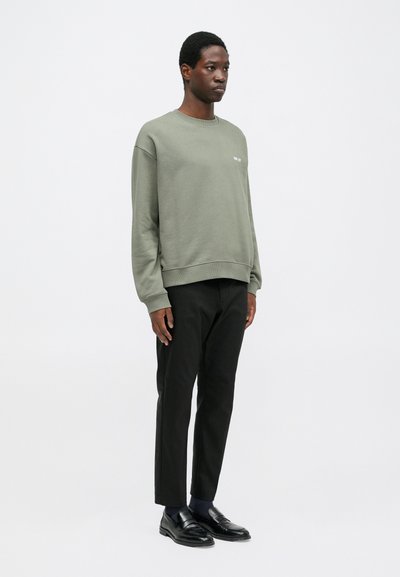 Olijfgroene sweatshirt met ribgebreide manchetten en onderkant, gecombineerd met zwarte broeken en zwarte instappers. Eenvoudig ontwerp met minimale branding.