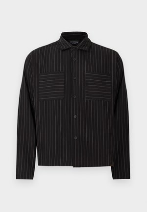 Čierna košeľa na gombíky s bielymi vertikálnymi pruhmi, s klasickým golierom a dvoma prednými vreckami. Vyrobená z hladkej tkaniny s voľným strihom.