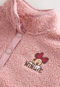Gros plan d'une veste polaire rose texturée avec des boutons-pression et un visage de Minnie Mouse brodé avec un nœud sur le devant.