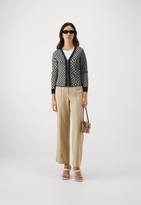 Svart-hvitt mønstret cardigan med V-hals, kombinert med beige vide bukser. Modellen holder en liten brun veske og har på solbriller.