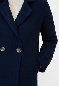 Marinblå dubbelknäppt kappa med ett strukturerat tyg, breda slag och två runda knappar framtill; sidofickor synliga.