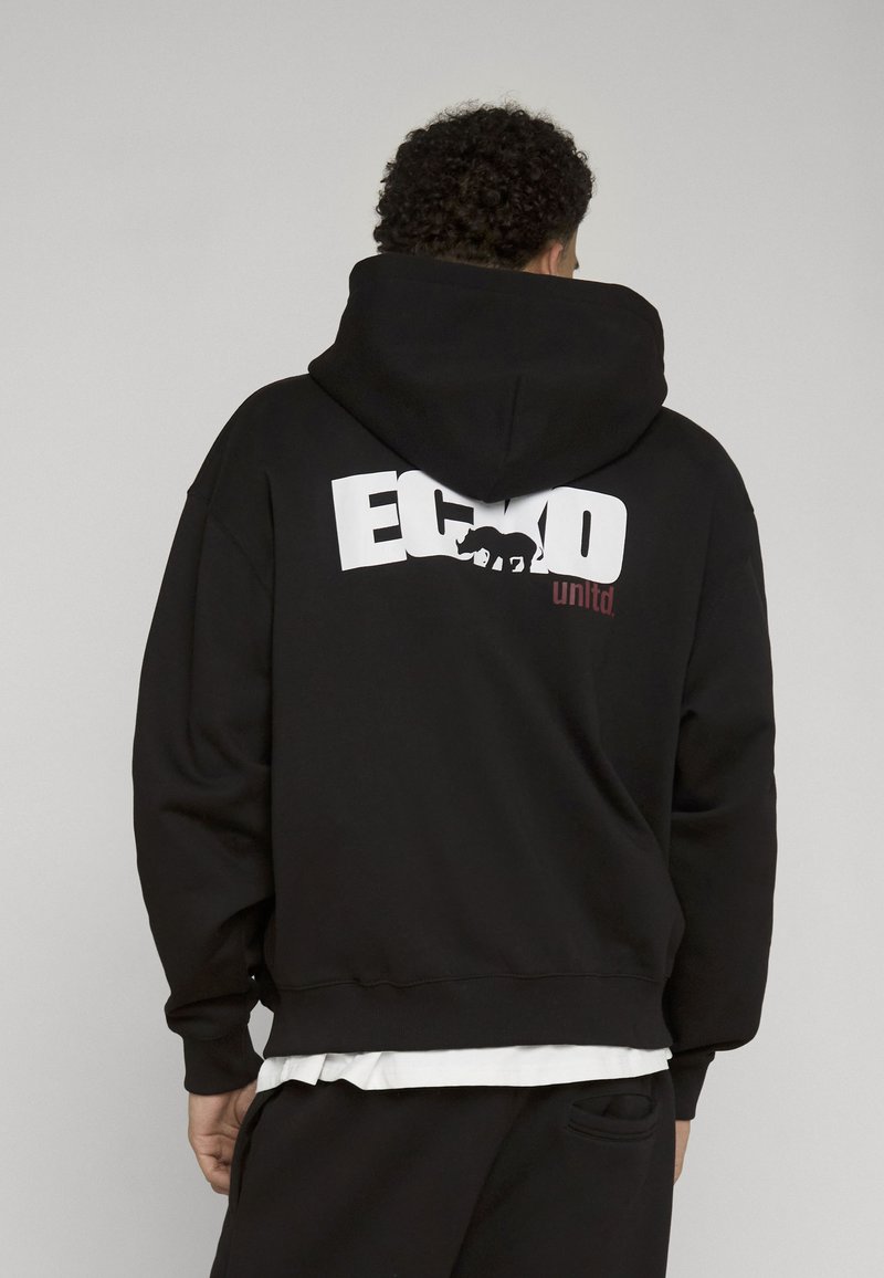 Personne aux cheveux bouclés, vue de dos, portant un sweat à capuche noir avec un grand "ECKO" blanc et un graphisme d'ours dans le dos, chemise blanche visible en dessous.