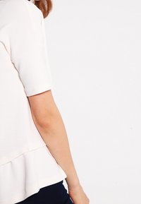 Vit kortärmad topp med peplumkjol, med en texturerad tyg. Den synliga armen är i en neutral miljö, vilket understryker plaggets design.