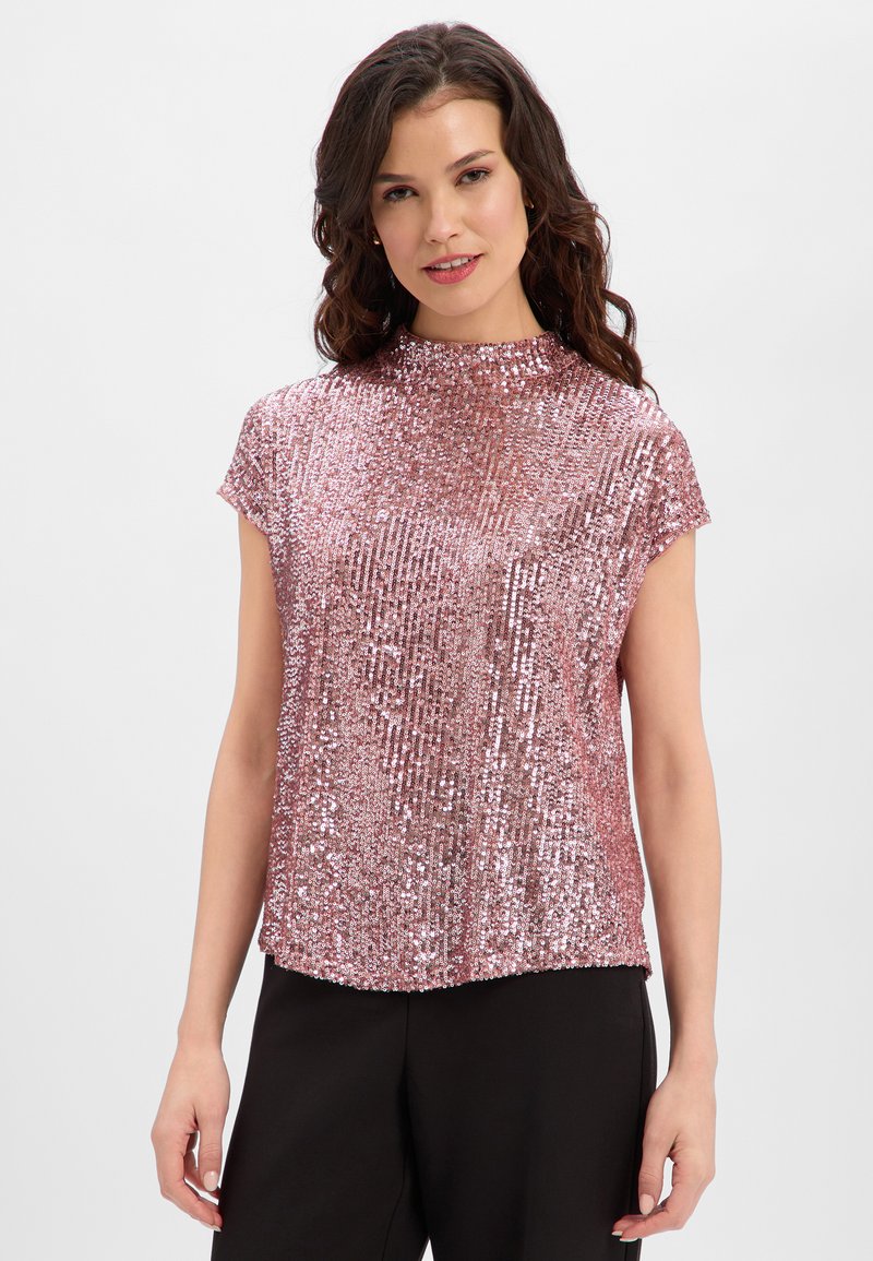 Woman with wavy brown hair wearing a shiny pink sequin short-sleeve top and black pants, standing against a white background.