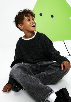 Grå strikket sweater med bred halsudskæring, båret over en hvid skjorte; grå denimbukser; siddende position med sorte fodtøj og en grøn abstrakt baggrund.