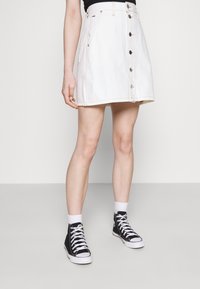 Gonna di jeans bianchi con bottoni frontali, tasche laterali e taglio dritto. Indossata con sneakers alte nere e calzini bianchi.