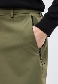 Χέρι μερικώς μέσα σε τσέπη με φερμουάρ σε χακί παντελόνι, που φοριέται με μαύρο μακρυμάνικο τοπ, αναδεικνύοντας τη λεπτομέρεια της τσέπης και τις ραφές.