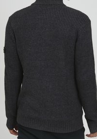 Pull en maille gris foncé avec un col haut, des poignets et un ourlet côtelés. Texture épaisse et accent de bouton sur la manche.