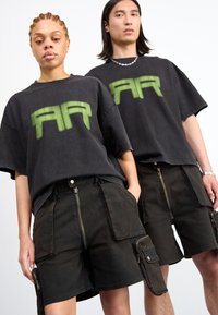 T-shirts oversized pretos com um logo verde "AA", combinados com calções cargo escuros com múltiplos fechos de correr e bolsos.